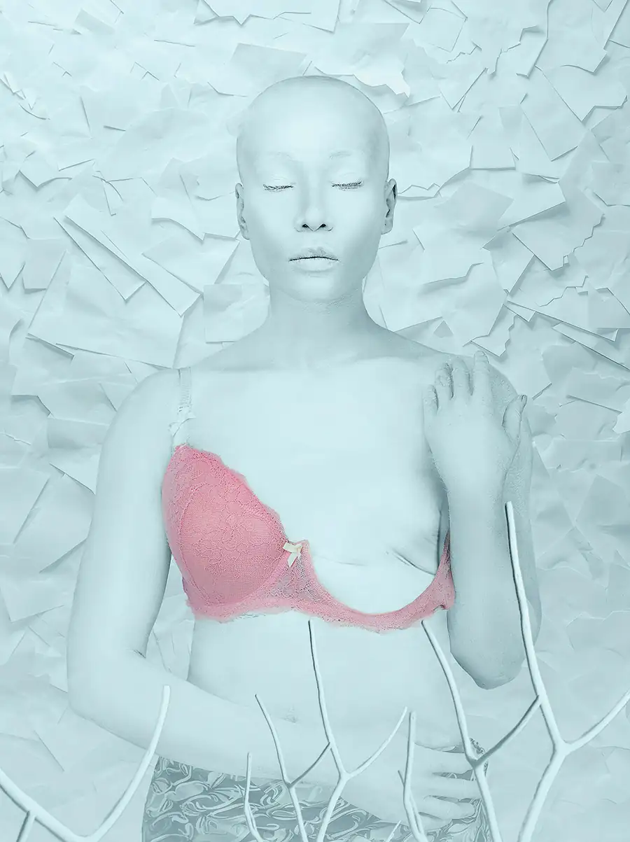 A bald woman with her eyes closed stands against a textured white backdrop, her skin painted pale and one pink bra cup visible, creating a powerful visual representation of cancer, vulnerability, and inner strength.