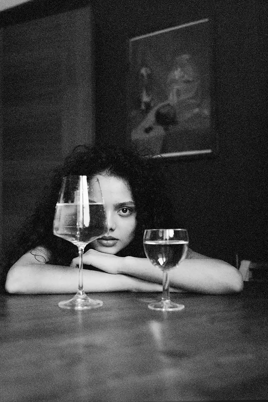 A woman in black and white resting her arms on a table and looking through two wine glasses, symbolizing how wine affects the skin and complexion.