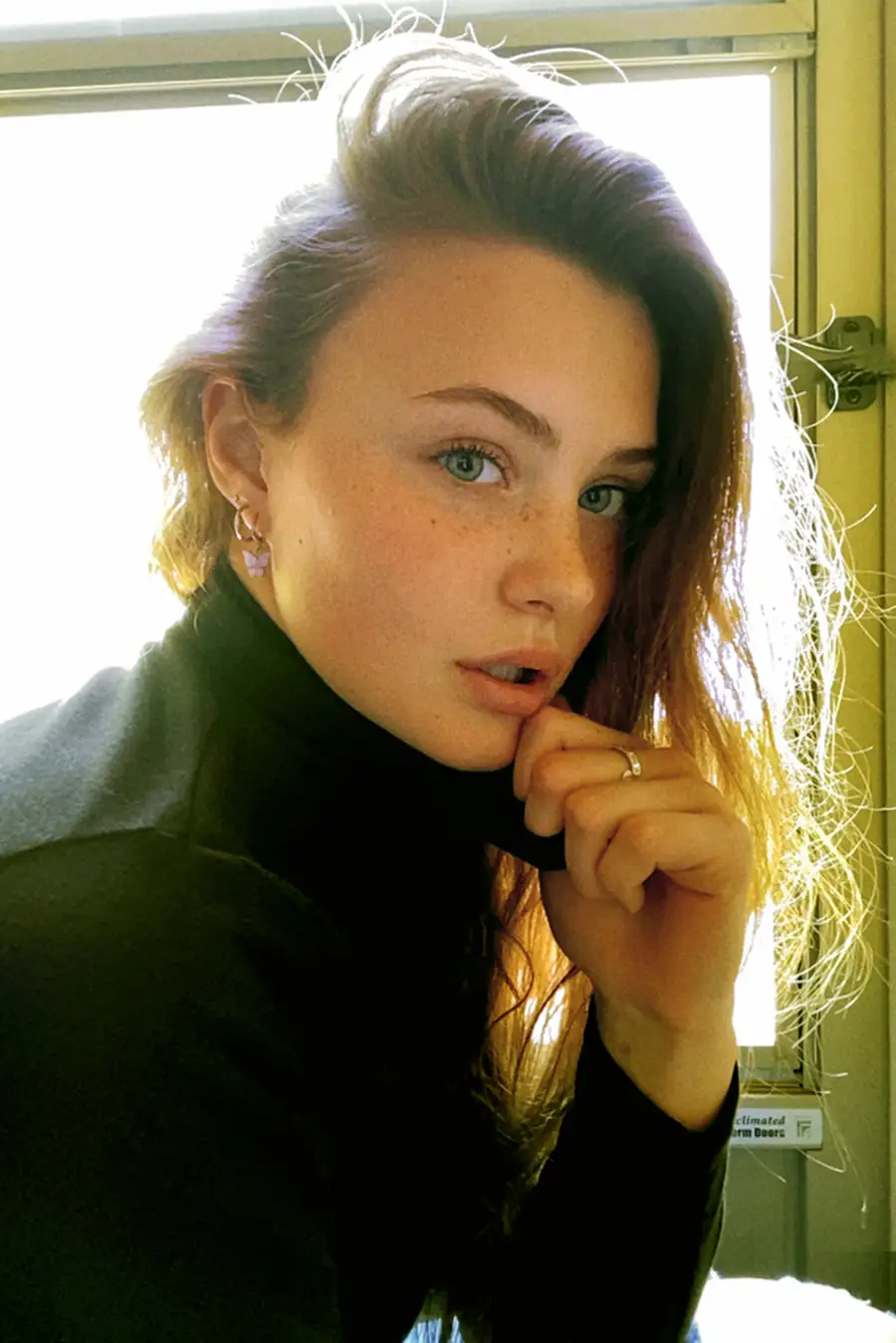 Bare Model Gibson in natural window light during a FaceTime photoshoot, freckles visible, minimal styling, unfiltered portrait.