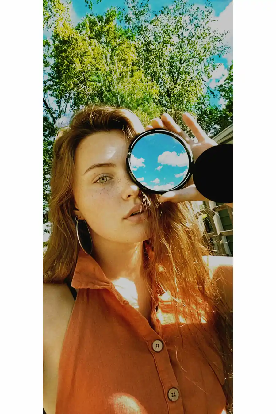 Bare Model Gibson during a FaceTime photoshoot, holding a camera lens to her eye with trees and sky in the background.