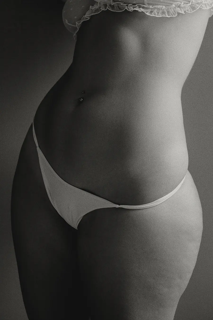 Black and white studio portrait of a woman’s torso showing natural curves, waist, and hip in soft window light.