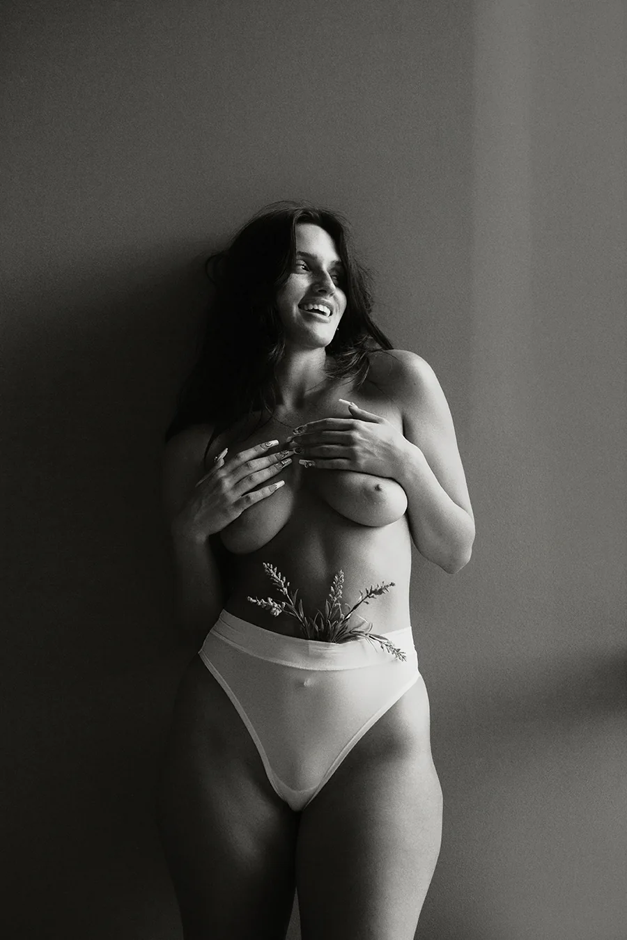 Black and white studio portrait of a woman standing naturally, holding flowers at the waist in soft window light.