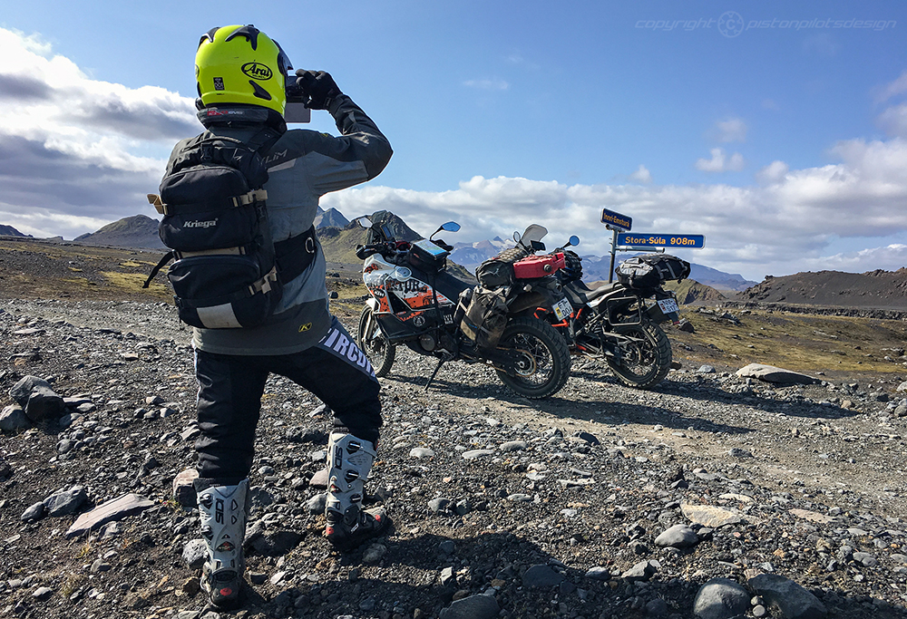 Person with bright yellow helmet photographing two adventure motorcycles on rocky terrain near a Stóra-Súla sign.