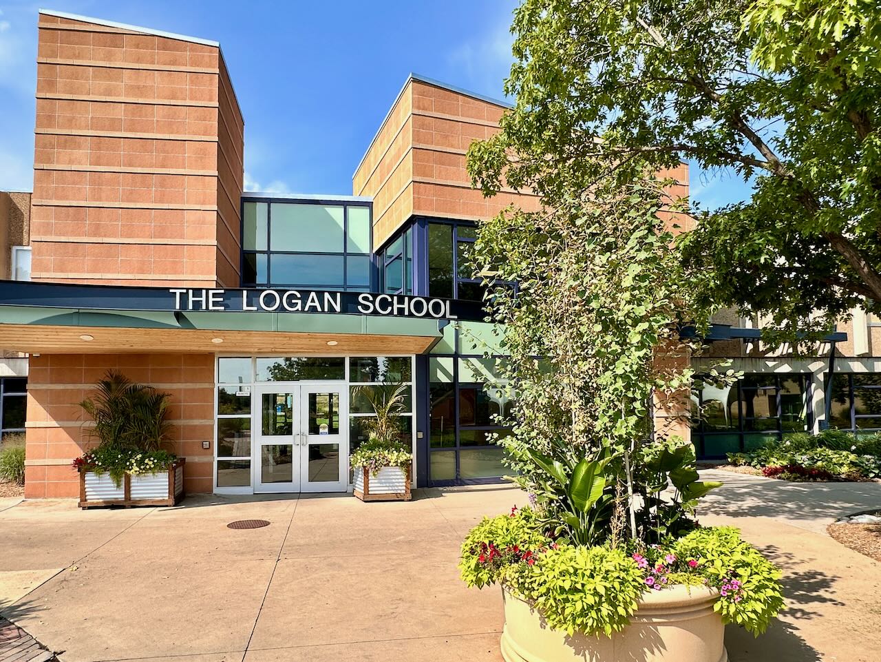 Exterior view of The Logan School building entrance on a sunny day, surrounded by green trees and flowering plants.