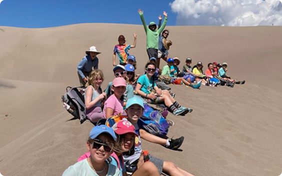 A group of students and teachers sitting and posing on a large sand dune under a blue sky during an outdoor field trip.