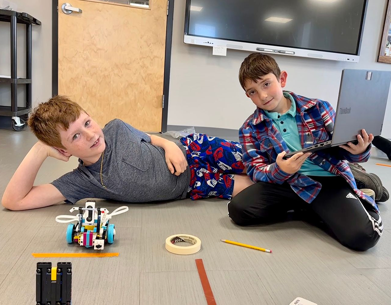 Two boys sitting on a classroom floor working together on a LEGO robotics project, with one holding a tablet and the other lying beside the assembled robot.