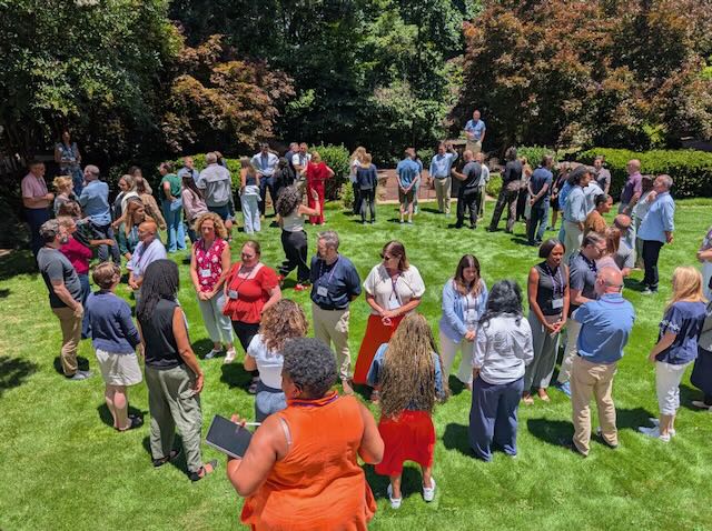 A group of adults standing in a circle on green grass with trees in the backdrop.