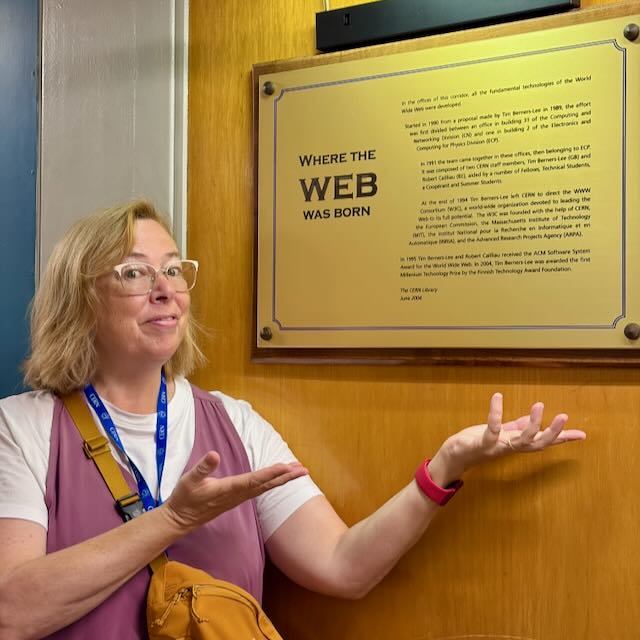 White adult woman with glasses and blond hair stands in front of a plaque at CERN.