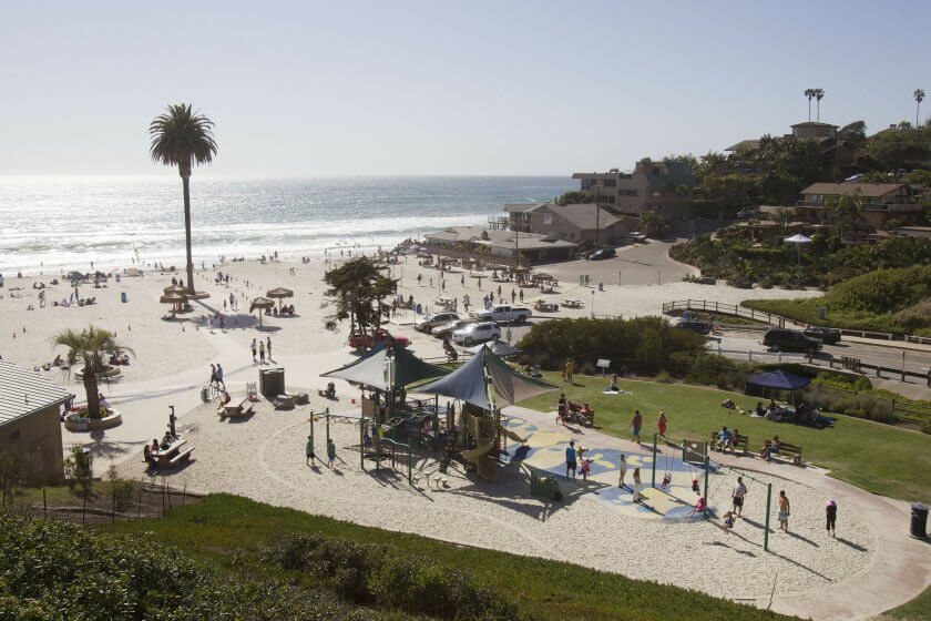 Wide sandy beach with a play structure and a parking lot to the right of it.