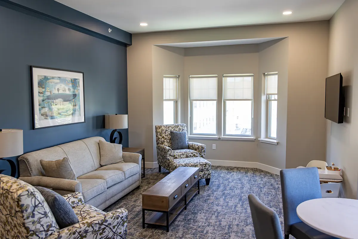 The living area of a room, complete with a couch, chairs, end table and TV. Bay windows have a view of the area around the house.