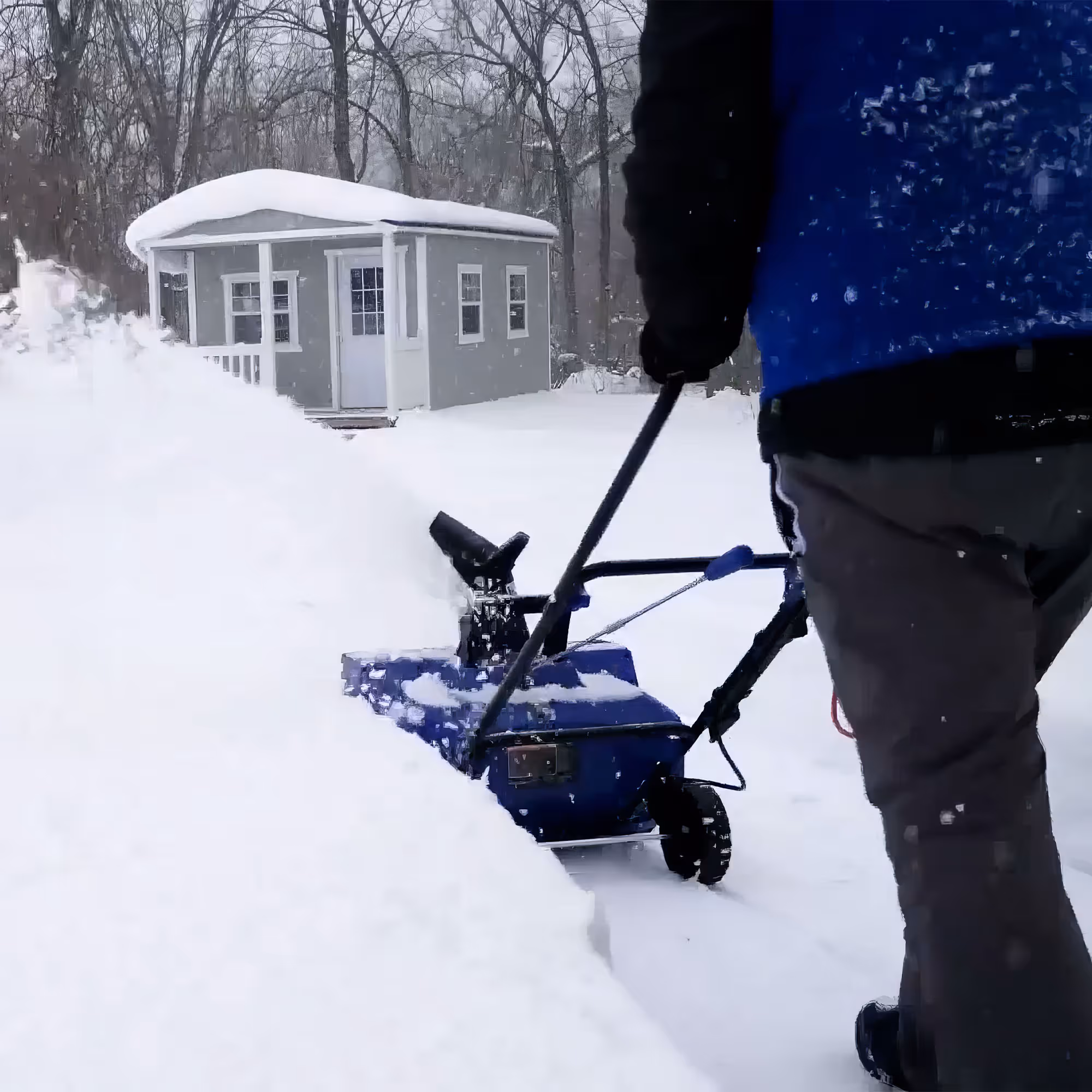 Person using a blue snow blower to clear snow outside a gray shed in a snowy wooded area.