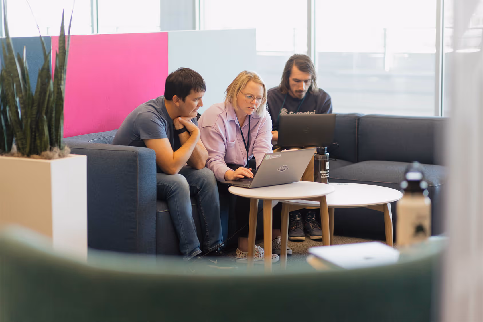 Three adults working on laptops together in a modern office lounge with a pink and blue divider.