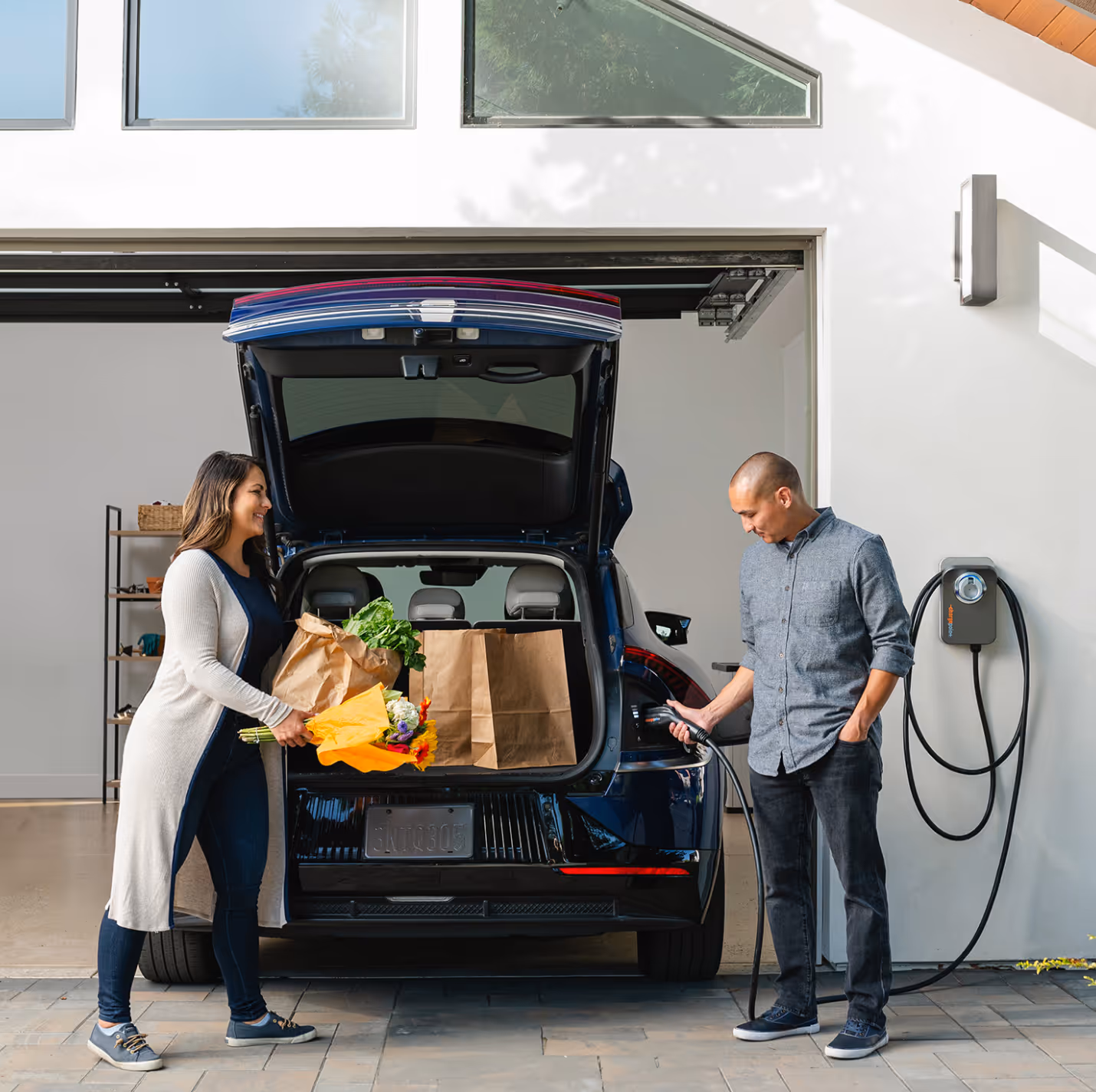 Man plugs electric car into wall charger while woman loads grocery bags and flowers into trunk.