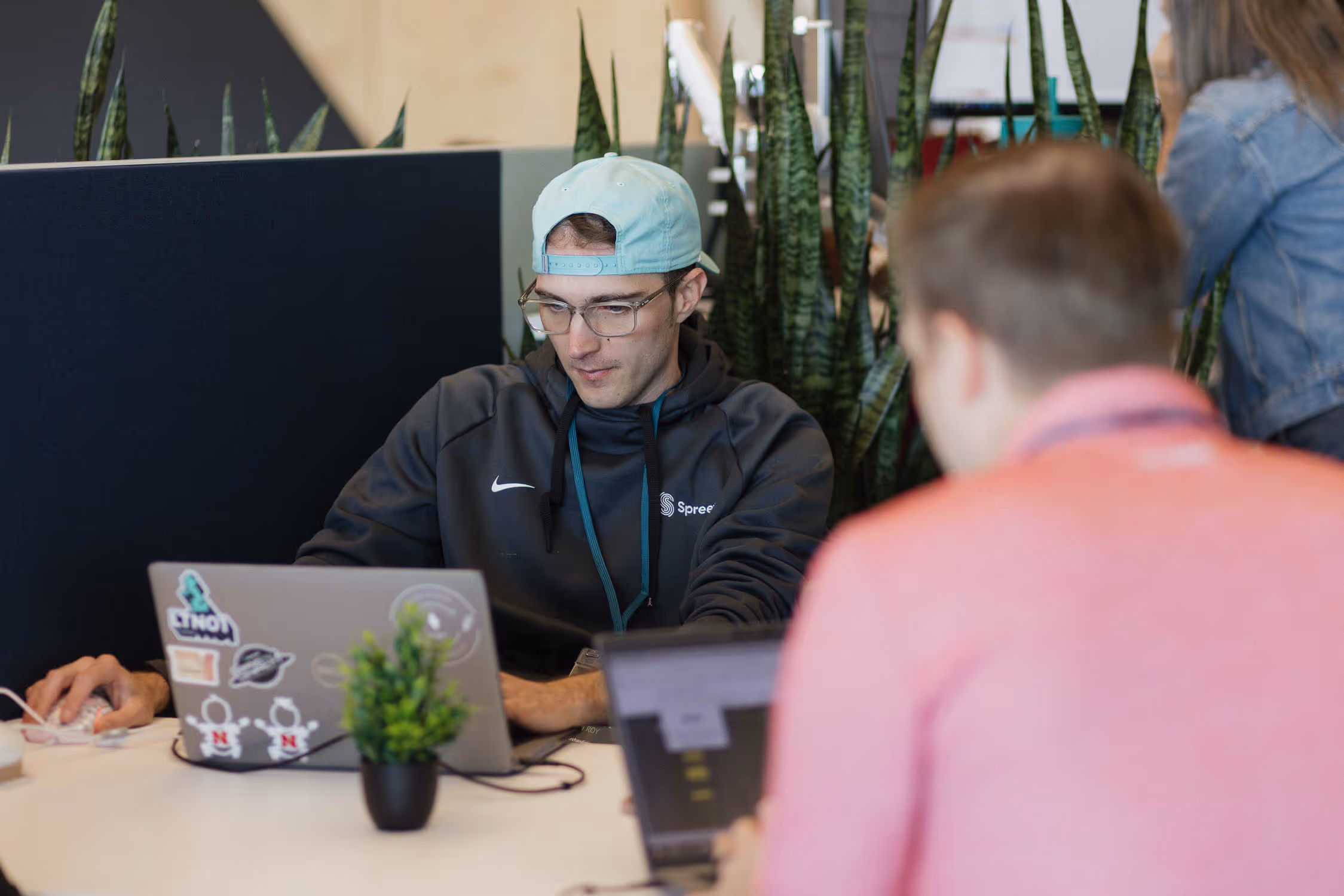 Young man wearing glasses and a cap works on a laptop with stickers in an indoor office with plants.