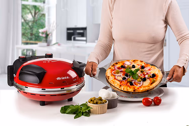 Person holding a freshly baked pizza next to a red Ariete pizza oven on a white kitchen counter.