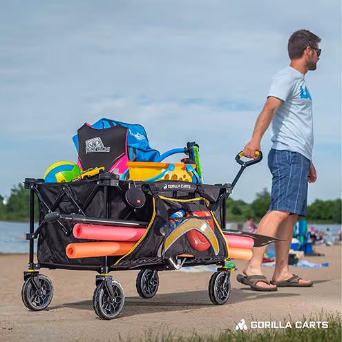 Man pulling a loaded Gorilla Carts wagon on a lakeside sandy beach with toys and gear inside.
