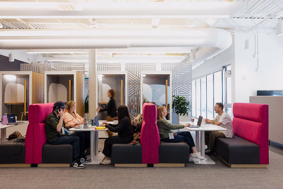 Indoor office space with several people seated in a long booth with bright pink high-backed seats working on laptops, talking, and using phones, with private phone booths in the background and a person walking by.