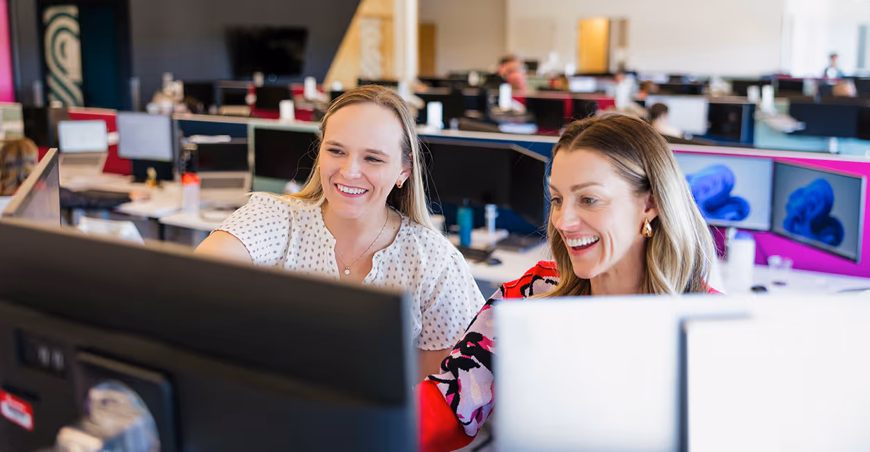 Two smiling women working together at a computer in an open-plan office with multiple desks.