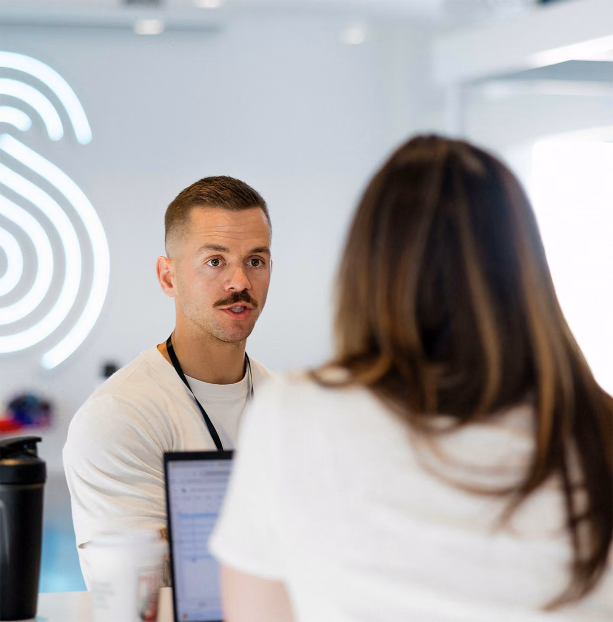 Man with mustache speaking to woman with long hair in a bright office space.