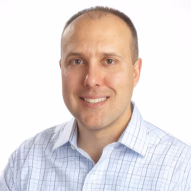 Smiling man with short hair wearing a light blue checkered shirt against white background.