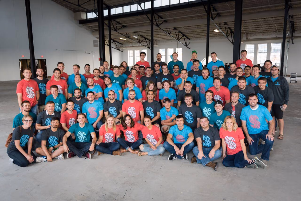 Group of people wearing red, blue, and gray T-shirts with logos posing in a large open indoor space.