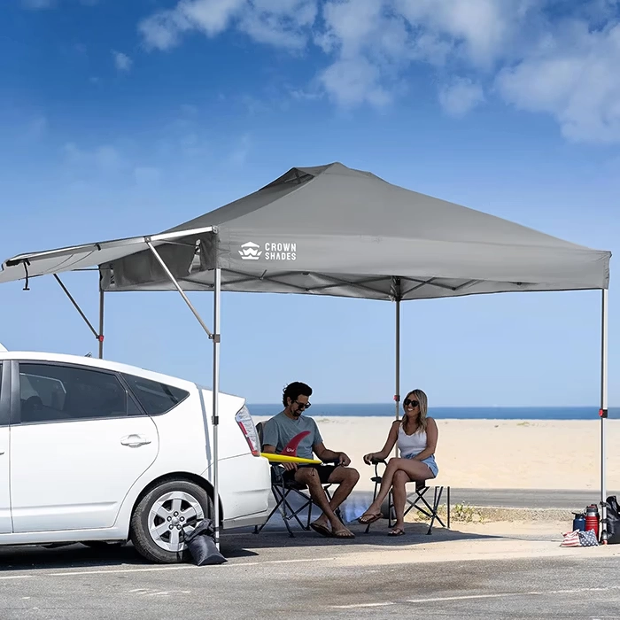 Man and woman sitting under a gray Crown Shades canopy near a white car on a sunny beach day.