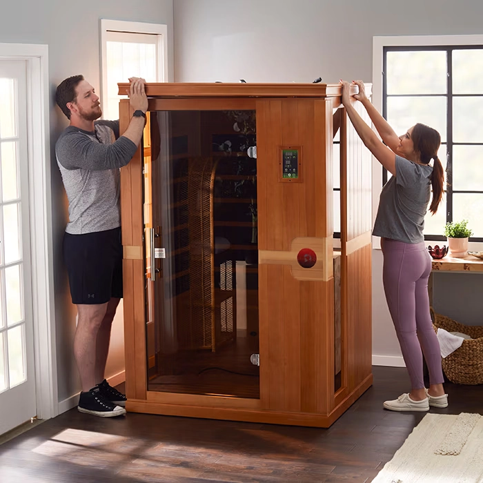 Man and woman installing a wooden infrared sauna cabin in a bright room with large windows.