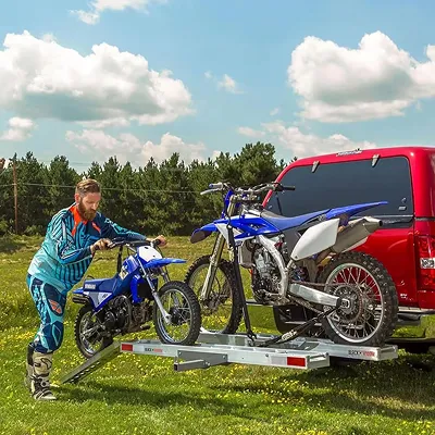 Man loading a small blue dirt bike onto a trailer carrying a larger blue dirt bike attached to a red vehicle.
