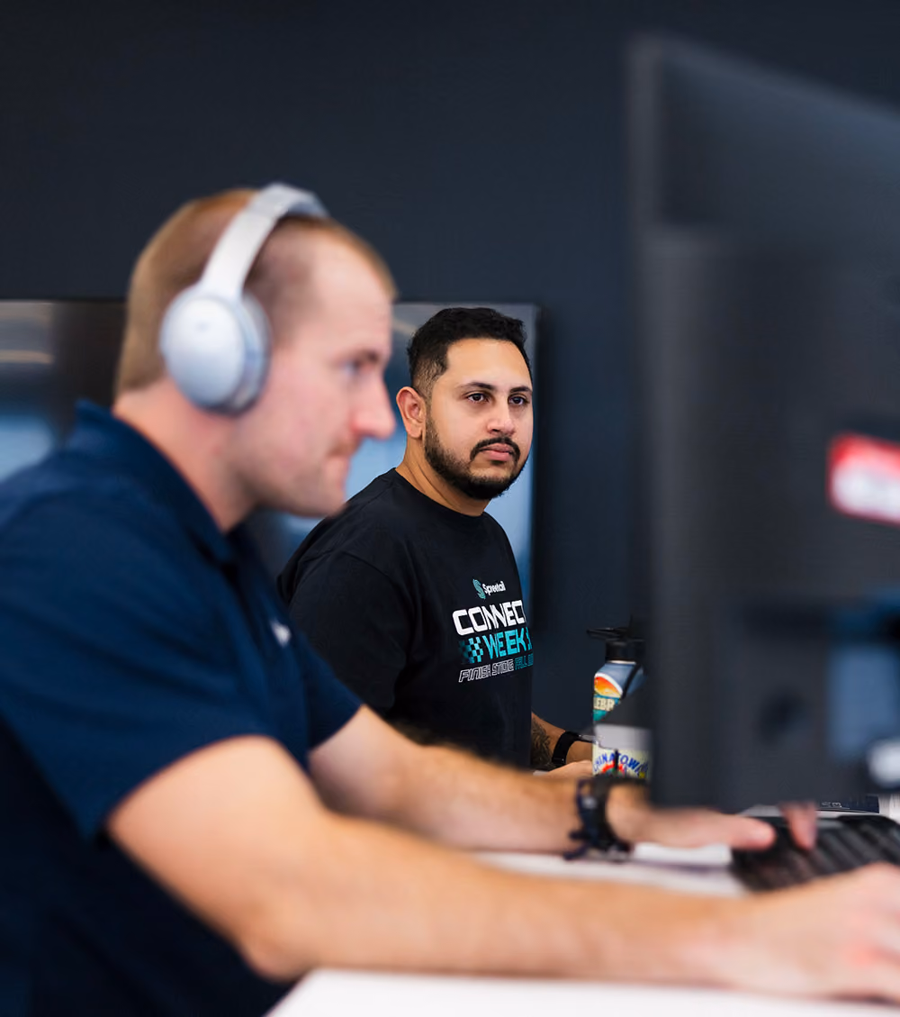 Two men working at desks with computer monitors; the man in the foreground wears headphones and the man in the background wears a black shirt with text and looks at his screen.