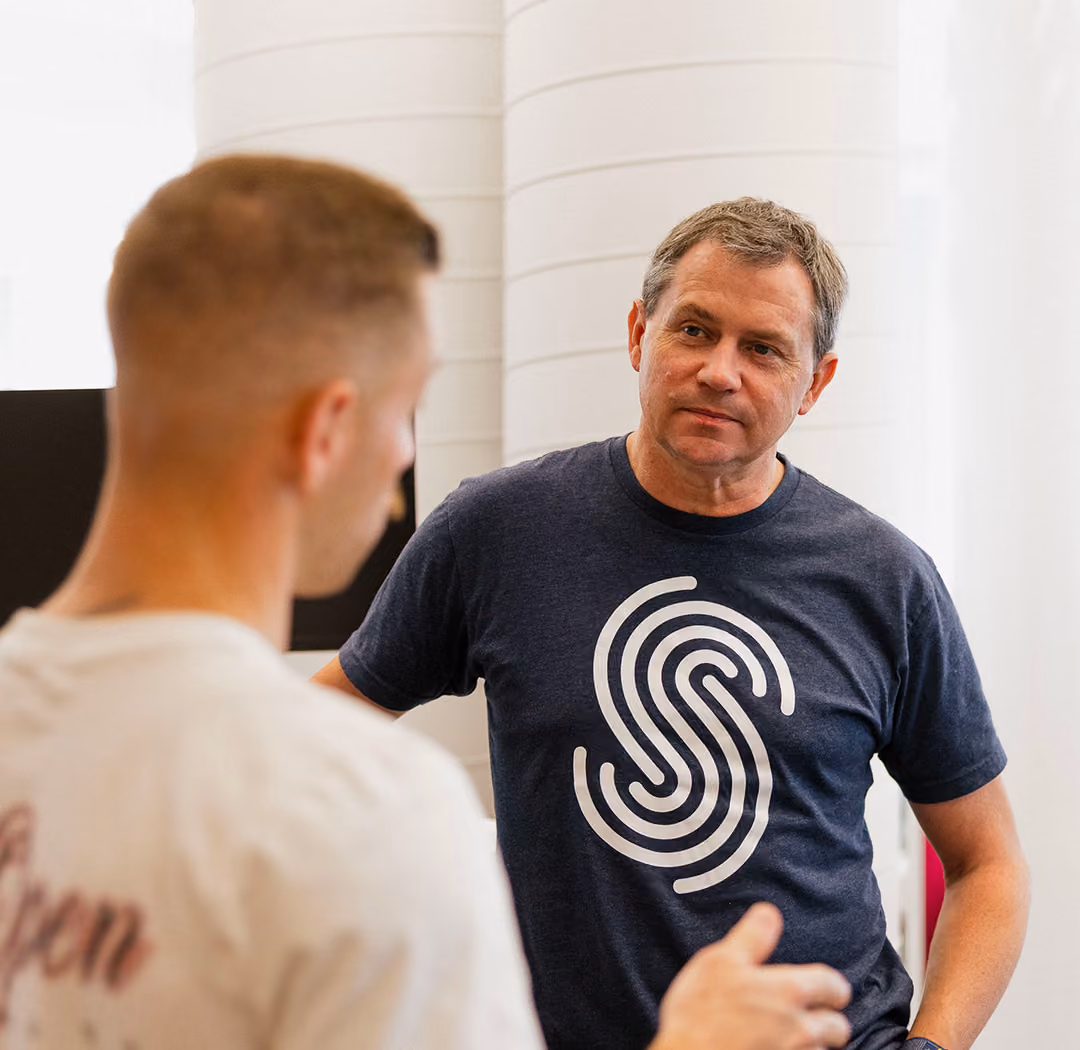 Two men engaged in conversation indoors, one facing the camera wearing a navy shirt with a large white stylized 'S' logo, the other shown from behind in a white shirt.
