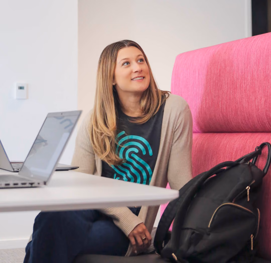 A woman with long blonde hair wearing a black shirt with a turquoise design and a beige cardigan sits next to a table with a laptop, looking up and smiling in a modern office setting with a pink cushioned chair and a black backpack beside her.