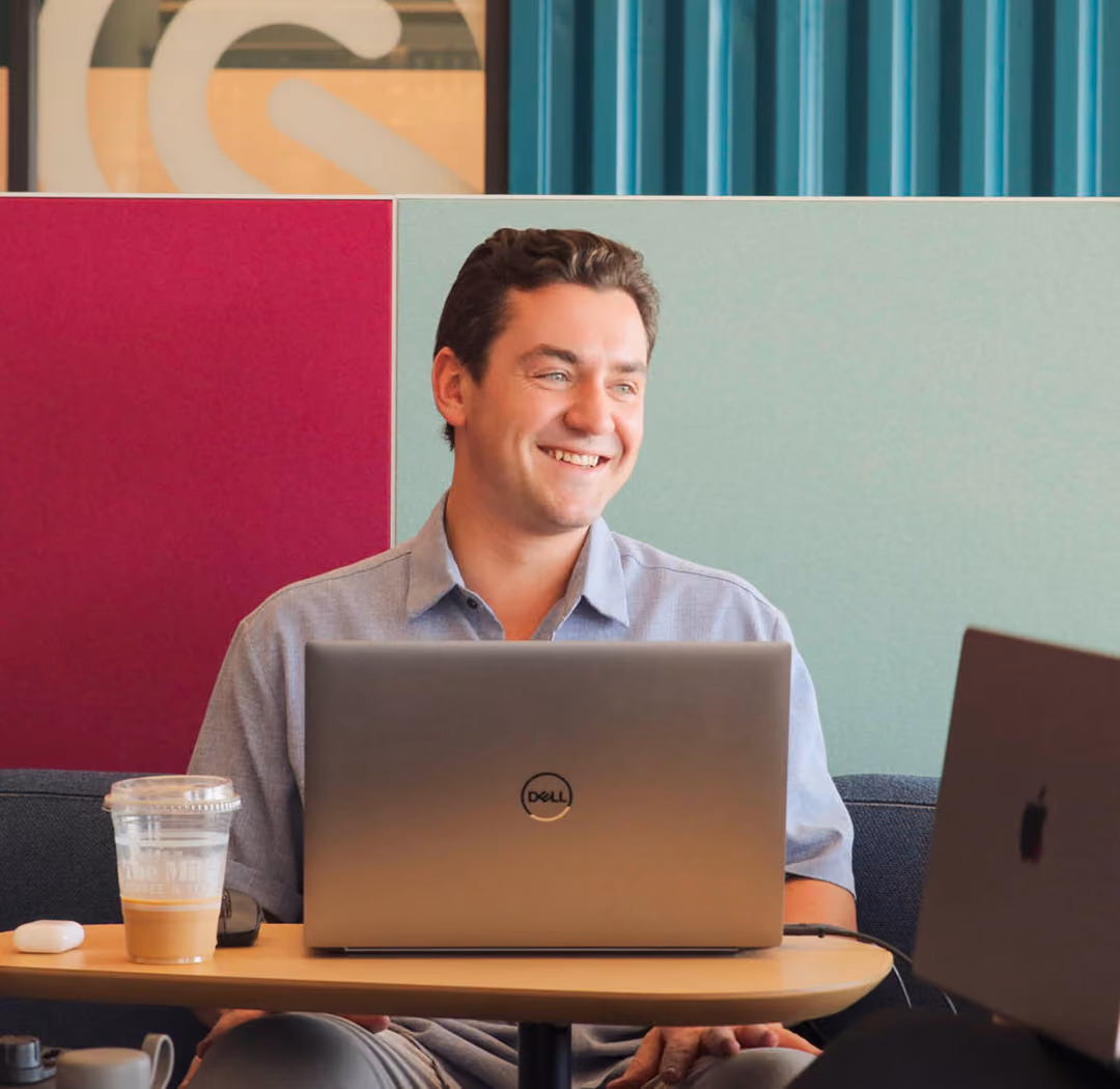 A smiling man sitting at a table with a Dell laptop in front of him and a cup of iced coffee beside it, with a colorful partition wall in the background.