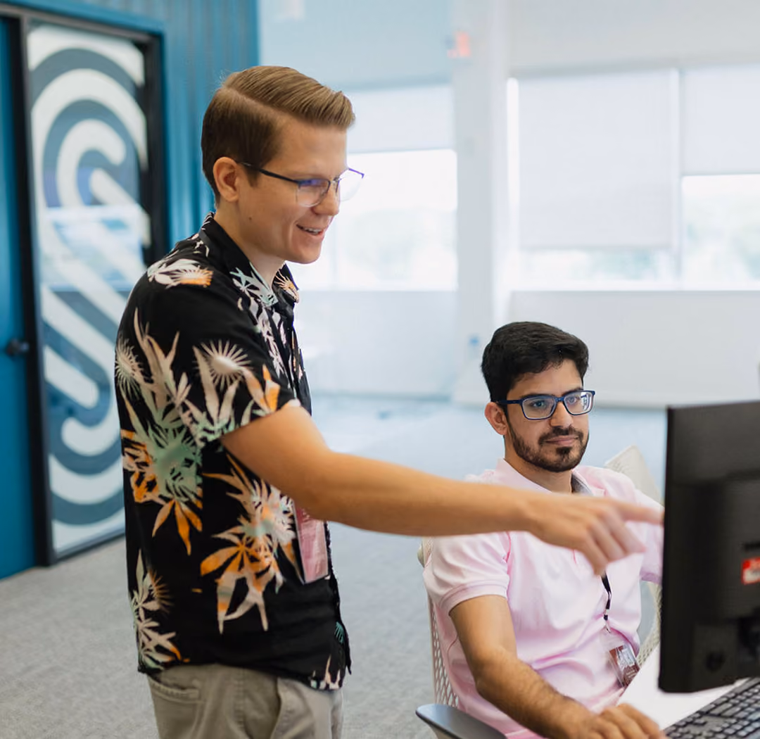 Two men with glasses collaborating in an office, one standing and pointing at a computer screen while the other sits and looks at the screen.