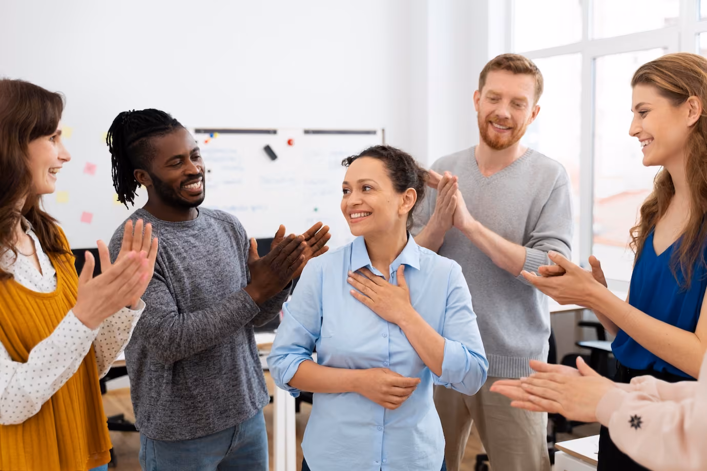 A new employee is smiling as she is being welcomed by her team of four co-workers surround her and are clapping for her.