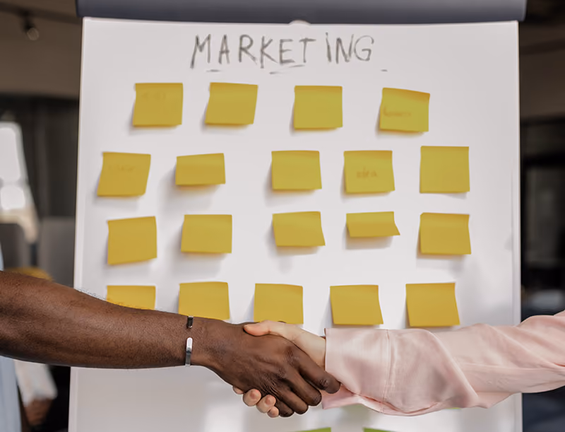 An employee shaking hands with their manager in front of a whiteboard showing a marketing plan after receiving positive feedback on their marketing strategy