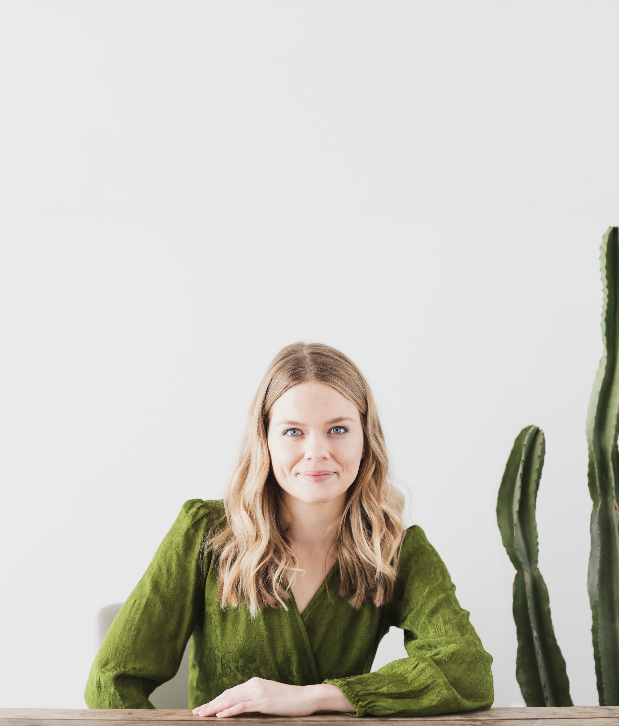 Shelby, a white woman with blonde hair wearing a green top, sits at a wooden table and smiles. There are two cacti in the background.