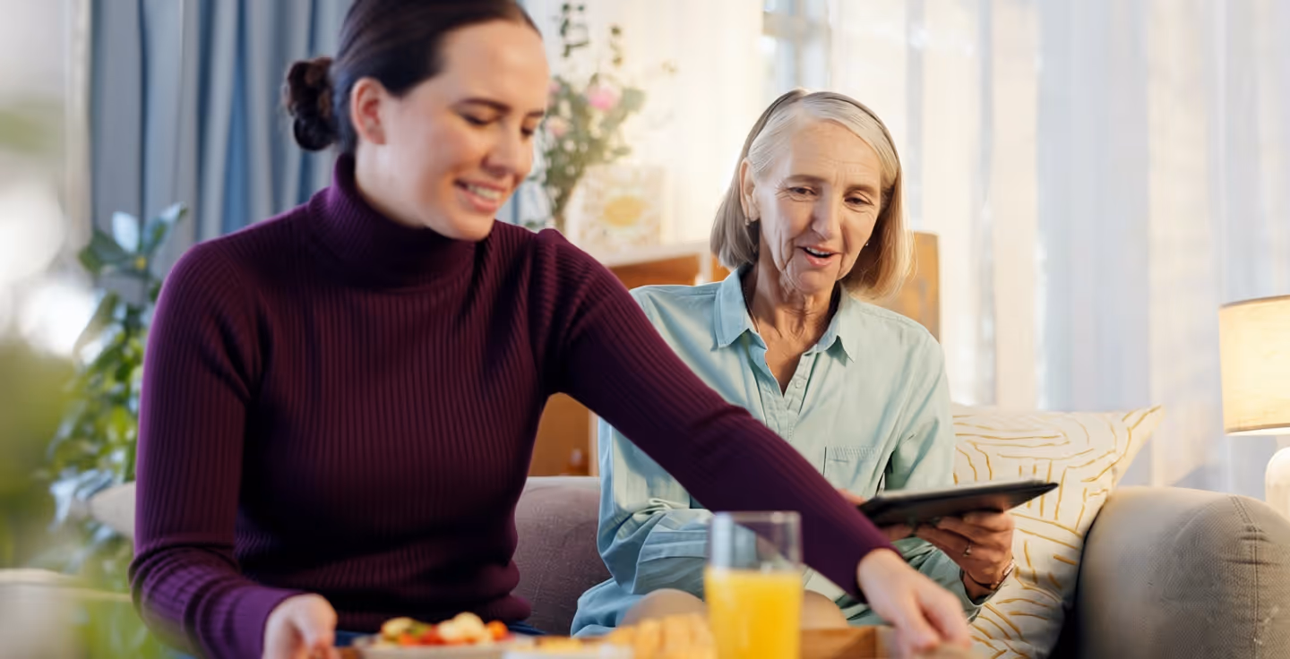 En kvinna serverar frukost till en äldre kvinna som sitter med en surfplatta.