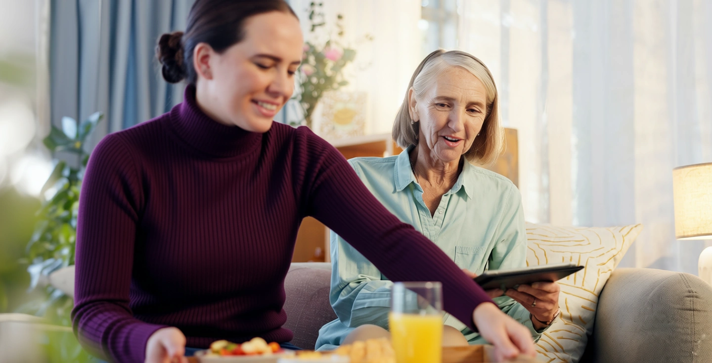 En kvinna serverar frukost till en äldre kvinna som sitter med en surfplatta.
