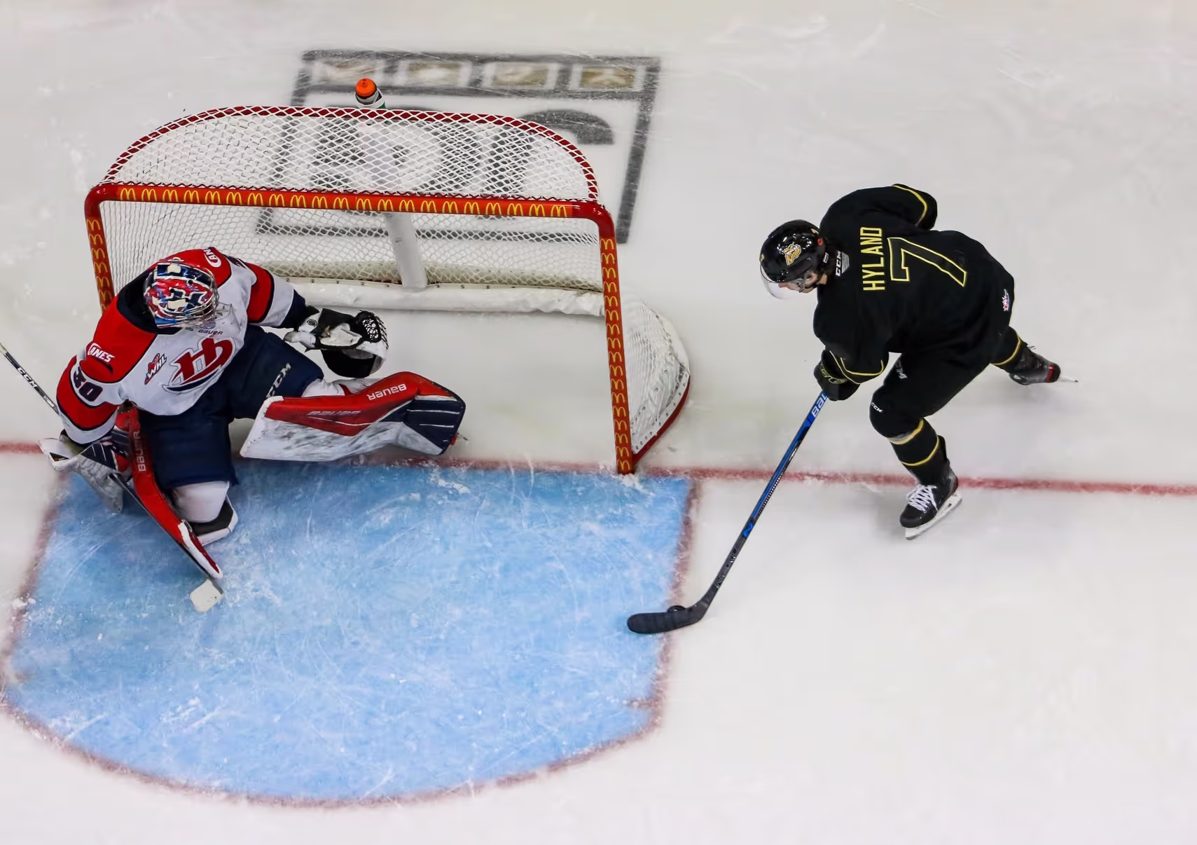 Hockey player in black jersey shooting the puck toward a goalie in white and red gear blocking the net.