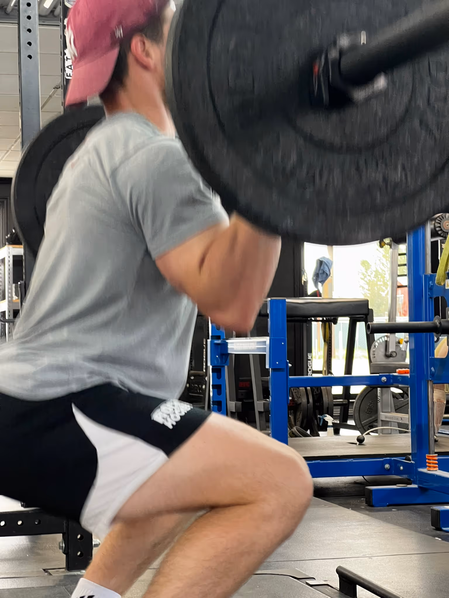 Man in a red cap and gray shirt performing a barbell squat in a gym.