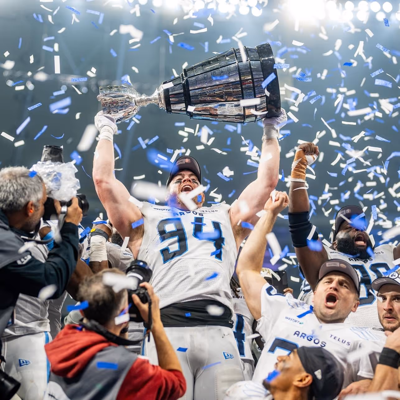 Excited Argos player #94 lifts trophy amid confetti with teammates and photographers celebrating.