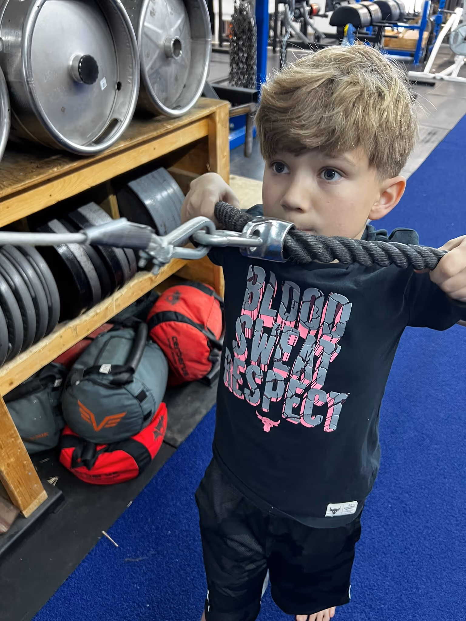 Young boy in a gym gripping a black rope attachment, wearing a black shirt with 'Blood Sweat Respect' text.