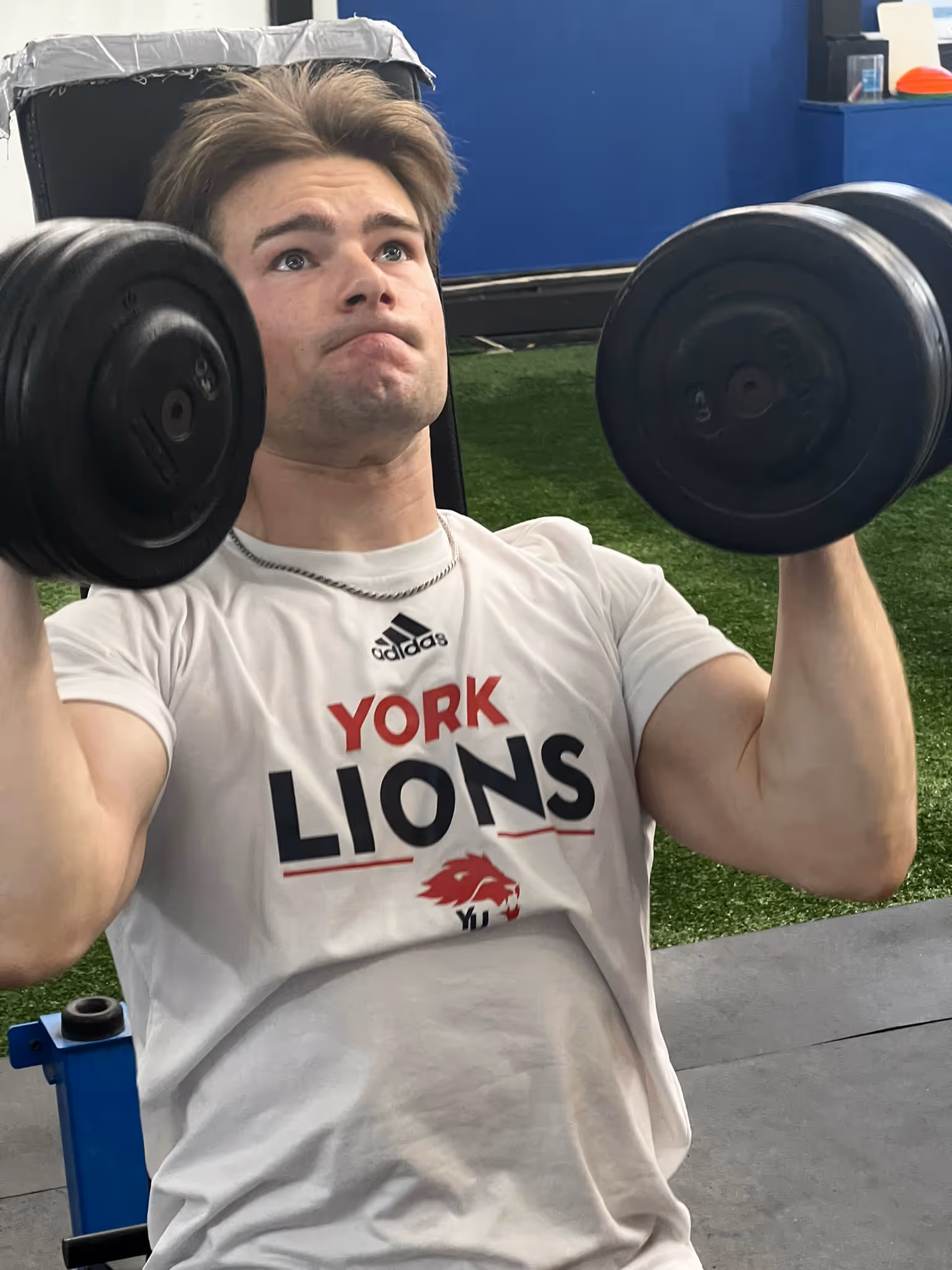 Young man wearing a York Lions shirt lifting black dumbbells in a gym setting.