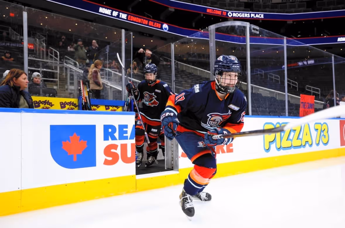 Youth ice hockey players in blue and orange uniforms skating onto the rink at Rogers Place arena.