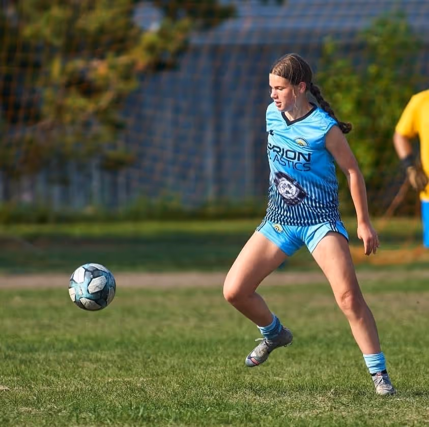 Female soccer player in blue uniform preparing to kick a ball on a grassy field.