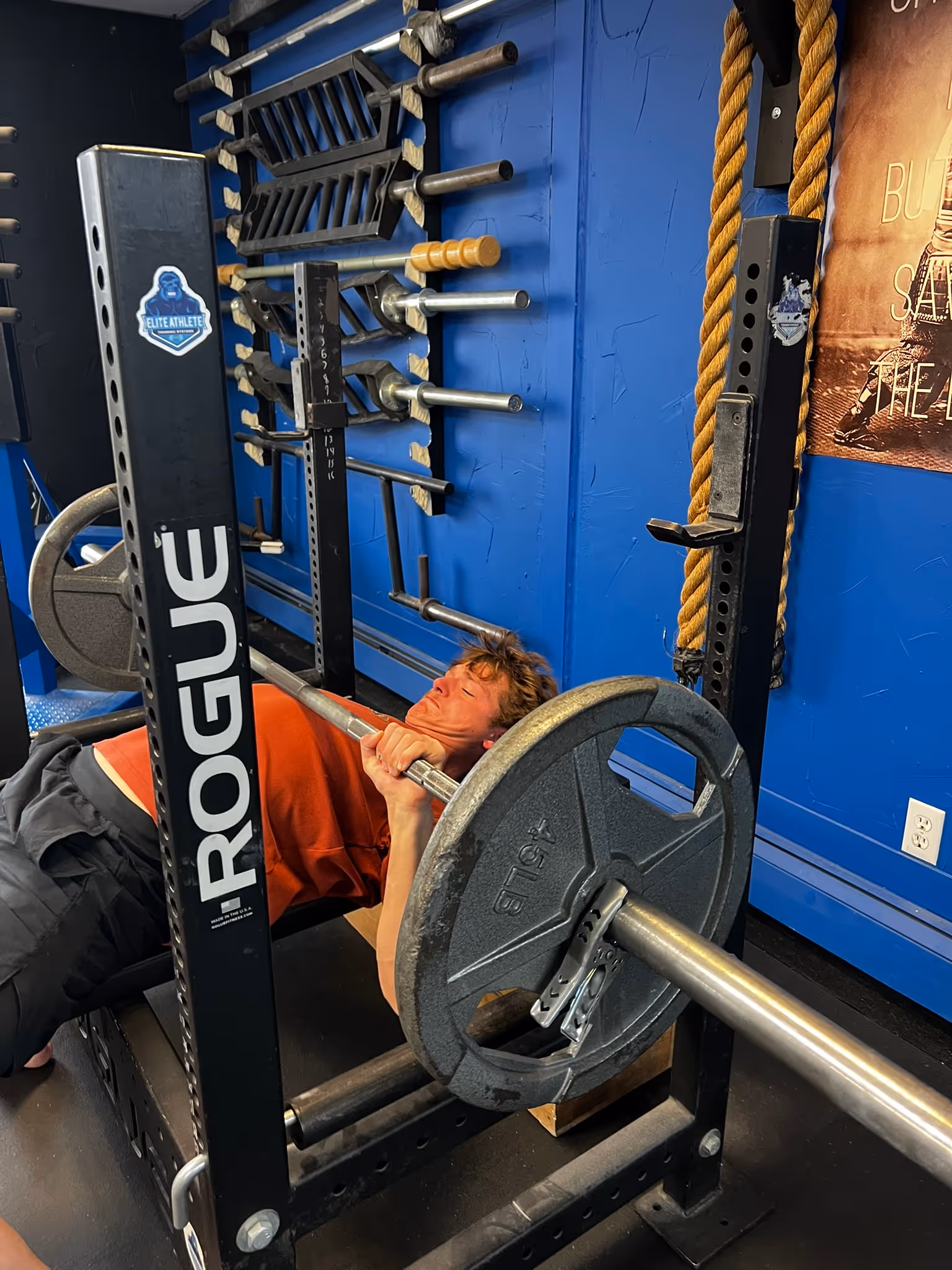 Person performing bench press with a barbell and 45 lb weight plates in a gym with blue walls.