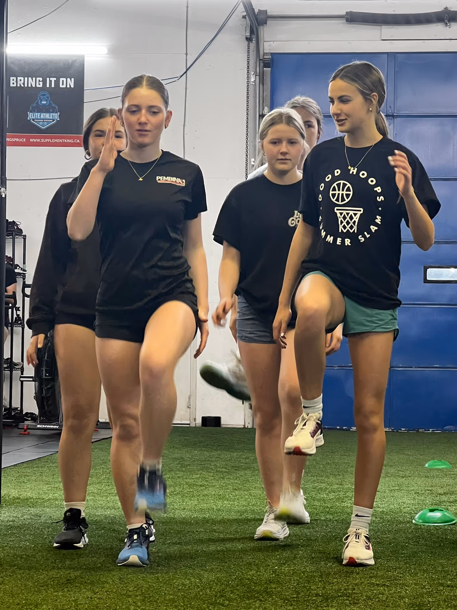 Group of young women exercising by lifting knees indoors on artificial turf with training cones nearby.