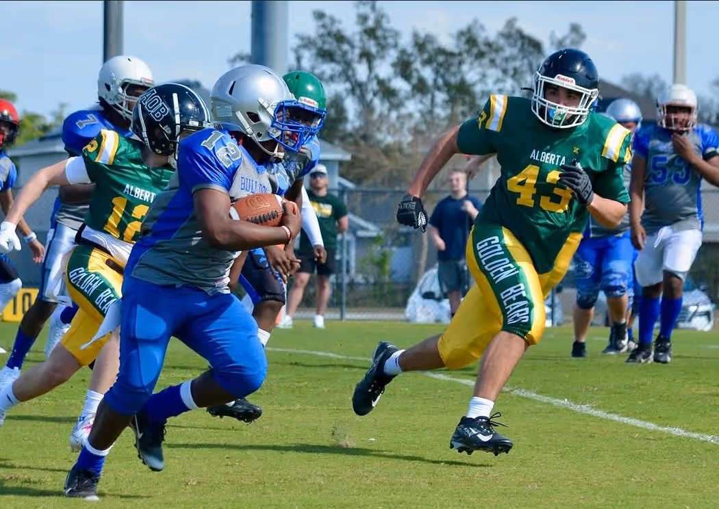 High school football player in blue and gray runs with ball while opponents in green and yellow chase him on a sunny field.