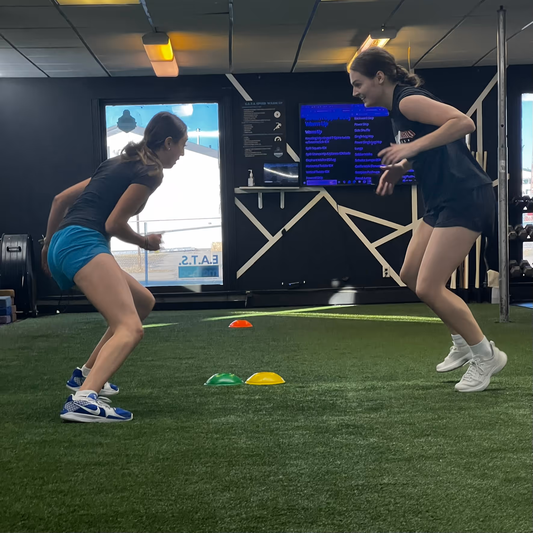 Two women in athletic clothes performing agility drills in an indoor gym with colored cones on turf.