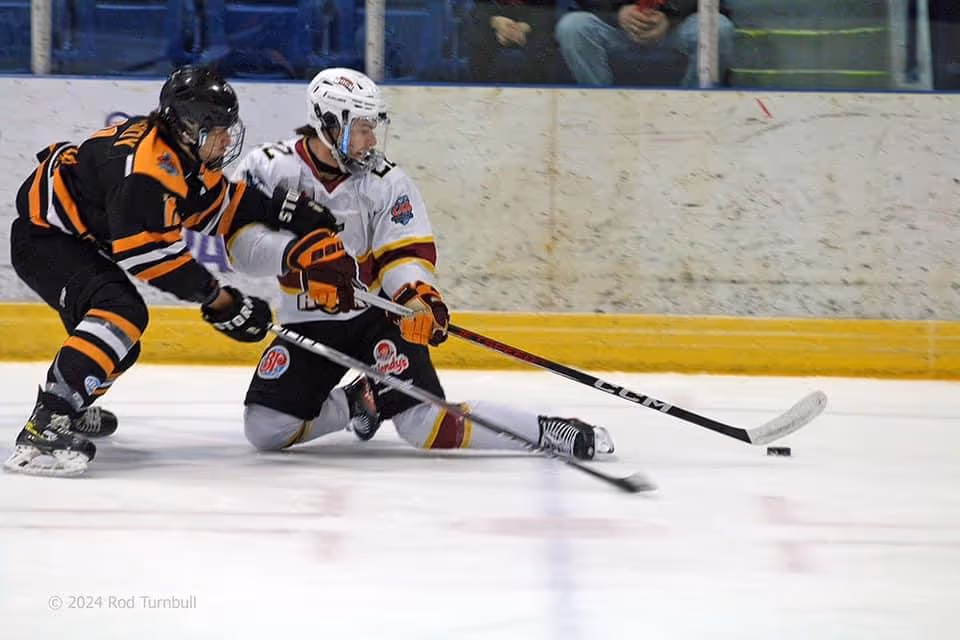 Two ice hockey players compete for the puck, one sliding on the ice while the other skates closely behind.